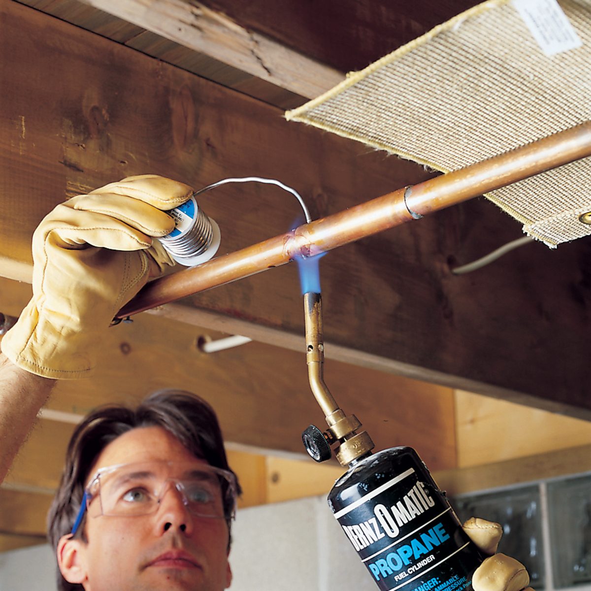 A person wearing safety glasses and gloves uses a propane torch to heat a copper pipe while applying soldering wire, performing a plumbing task under a wooden ceiling.