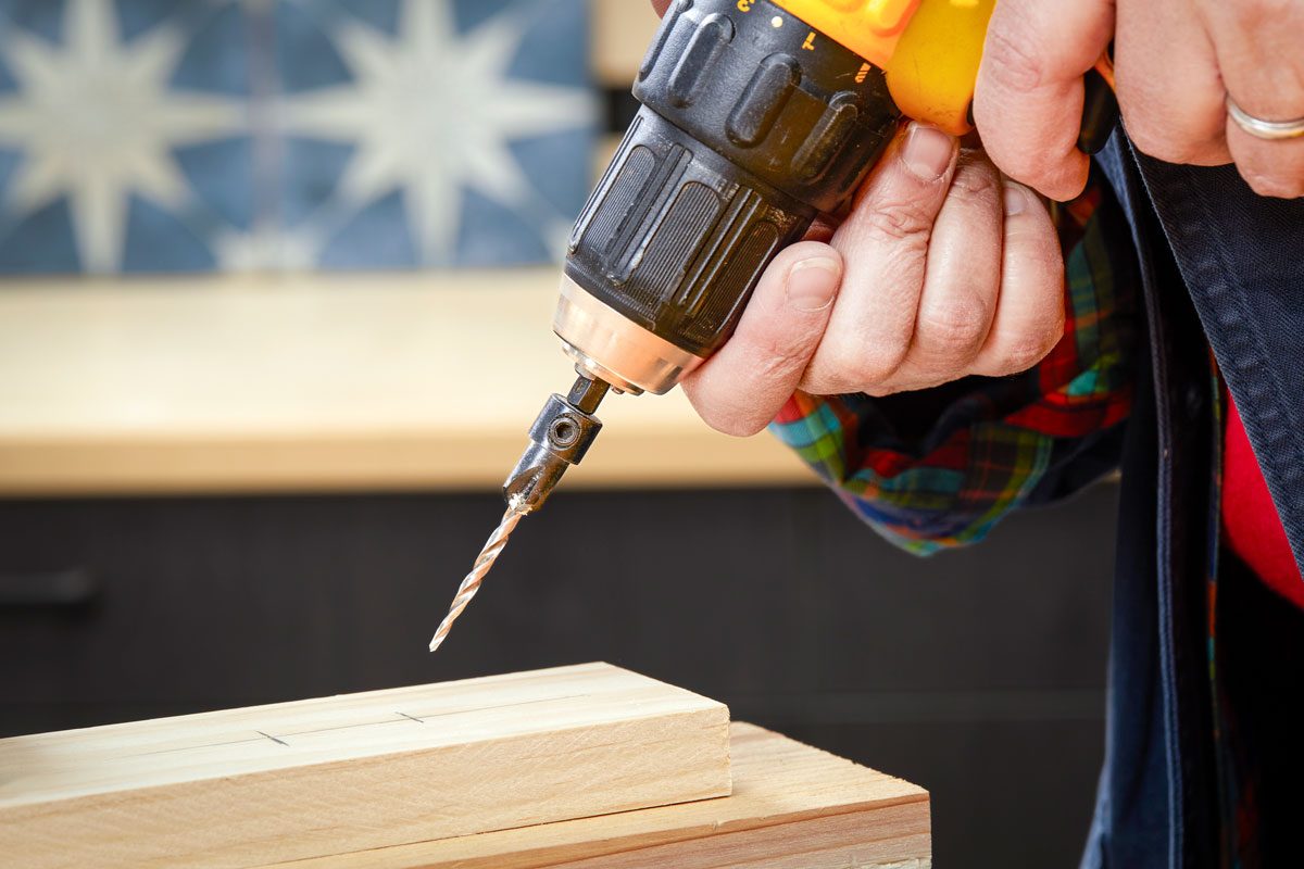 a close up view of a hand holding a yellow and black drill; a drill bit is inserted into the chuck and is positioned just above a piece of light colored wood; the background is blurred but appears to show a wooden surface and a tiled wall with a blue and white star pattern