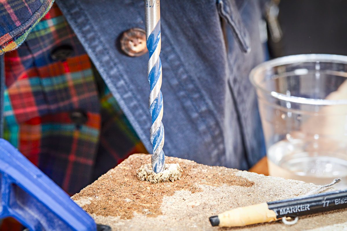 a drill bit, with blue and silver flutes, drilling into a light brown, granular material, to the left, a blue clamp is visible, a pencil lies to the right of the material, and a clear glass containing liquid is in the background