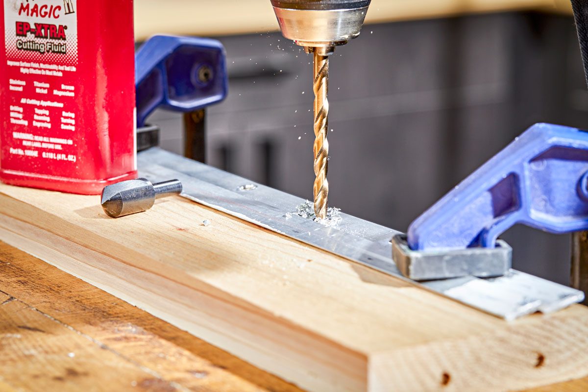 a drill bit, drilling through a piece of flat, silver colored metal; the metal is secured to a wooden surface with two blue clamps, one on each side; a small amount of metal shavings is visible around the drilling point; to the left of the metal, a red bottle is visible
