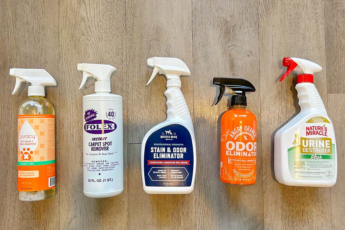 Five different pet stain and odor remover spray bottles are lined up on a light wood floor. Each bottle has distinct labels and spray nozzles in various colors.