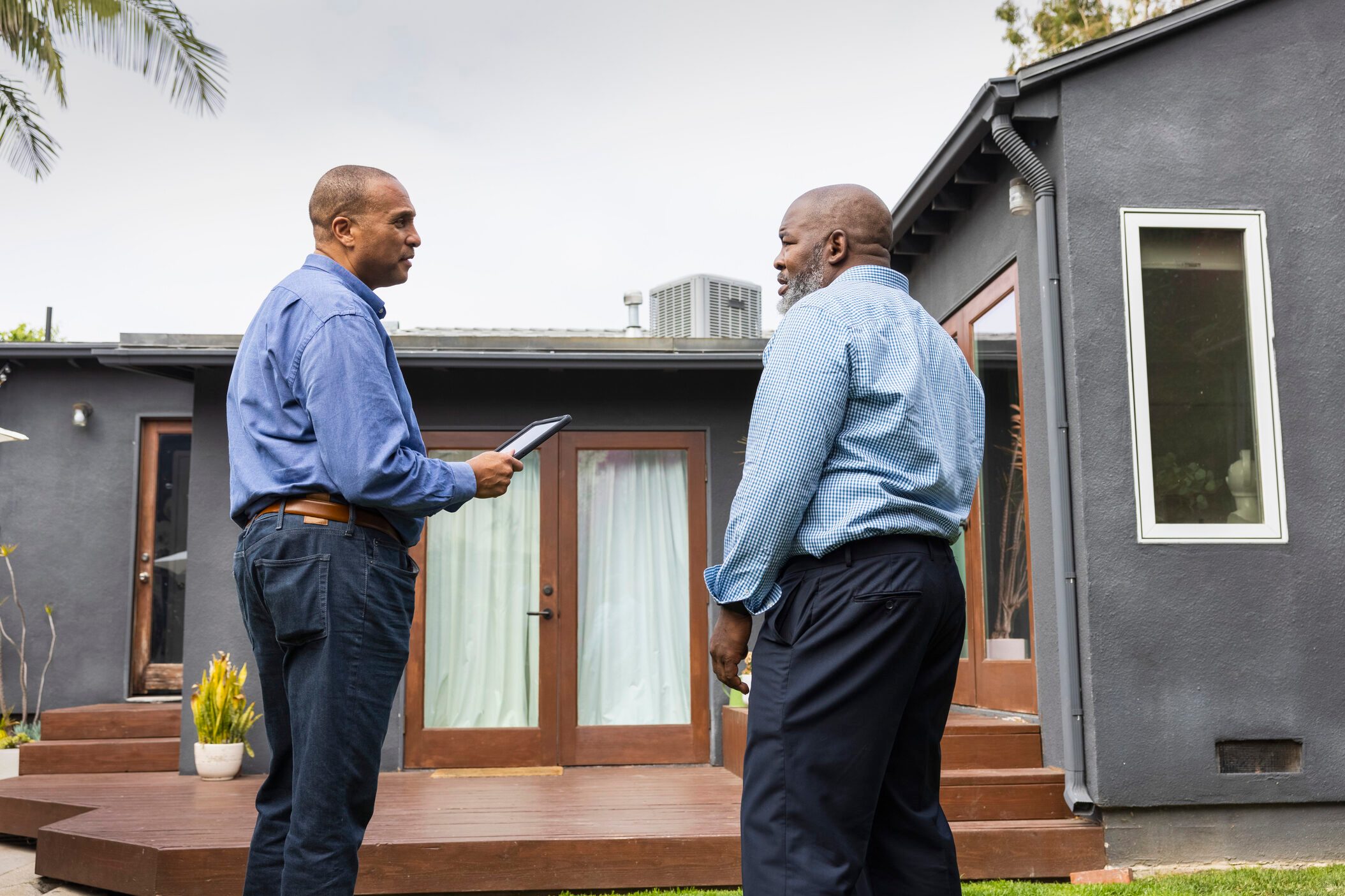 Mature salesman holding digital tablet while discussing with male customer outside house