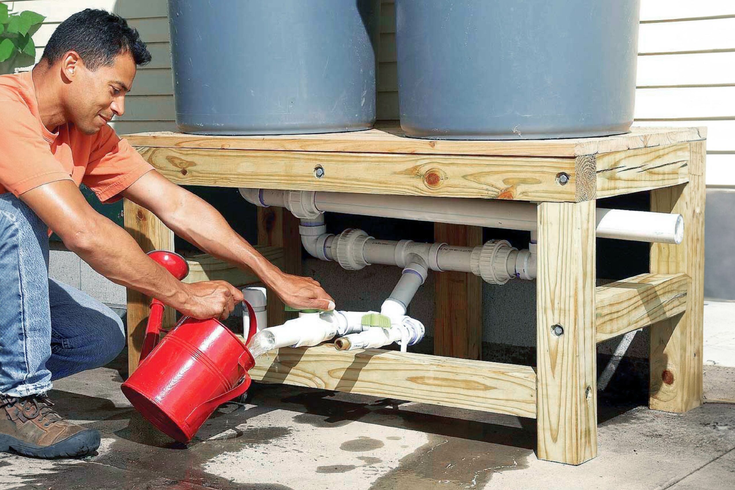 A man pours water from a red watering can into a pipe system connected to two gray barrels, surrounded by wooden framing and concrete flooring.