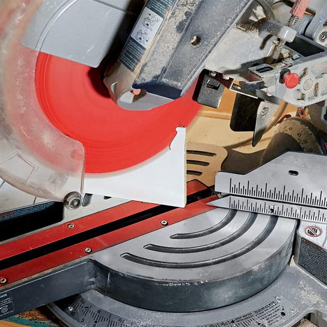 A close-up of a miter saw with a bright red circular blade, a metal guide fence, and measurement markings, set on a workbench ready for cutting materials.