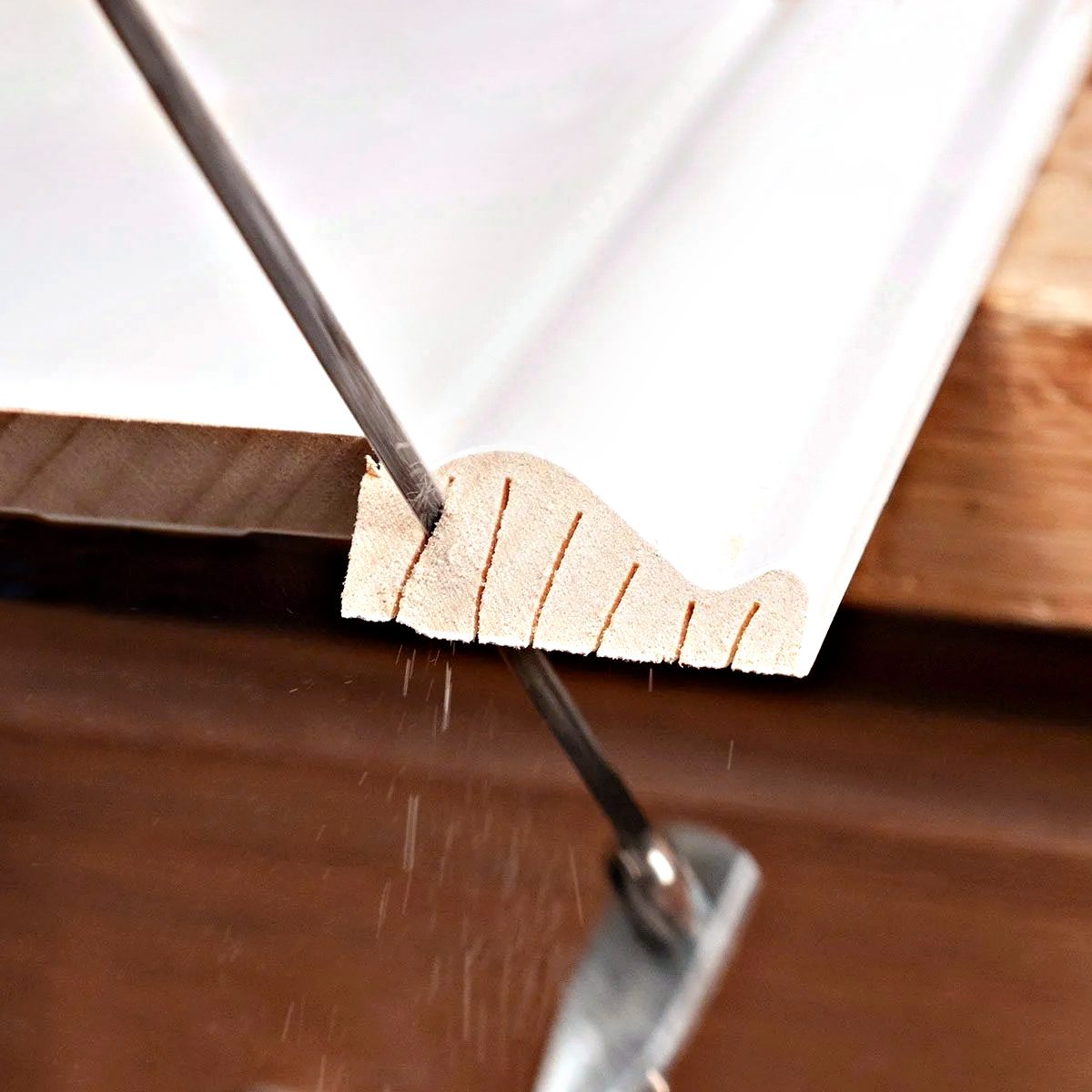 A hand saw is cutting through a piece of white trim molding, with several small kerf cuts visible along the angled edge. Sawdust is falling as the blade moves through the wood.