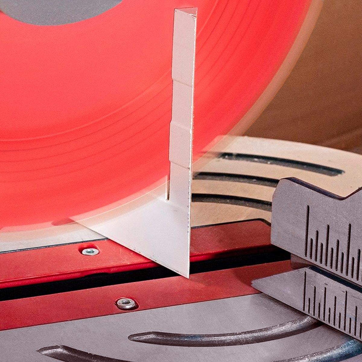 A close-up of a circular saw cutting through a white rectangular object, with a metal ruler nearby showing measurement markings. The saw blade is red and partially visible.