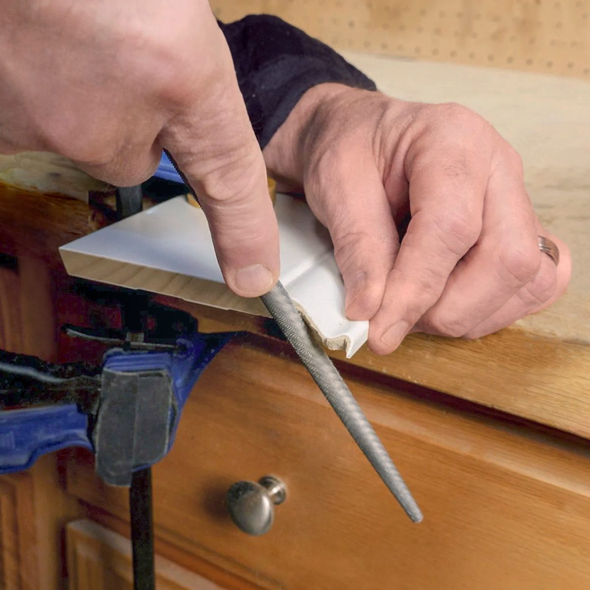 A person uses a round metal file to smooth the edge of a piece of wood clamped to a workbench.