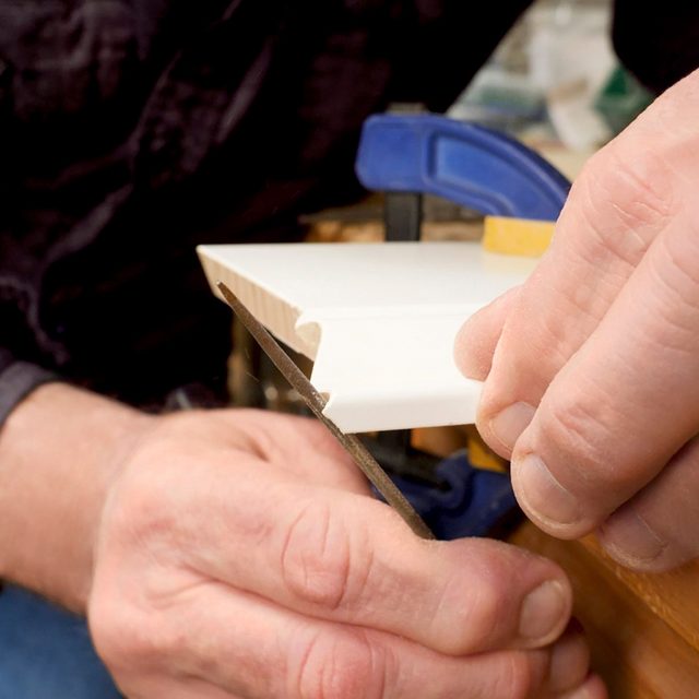 A person uses a hand saw to cut a piece of white trim or molding, which is held steady with a blue clamp.