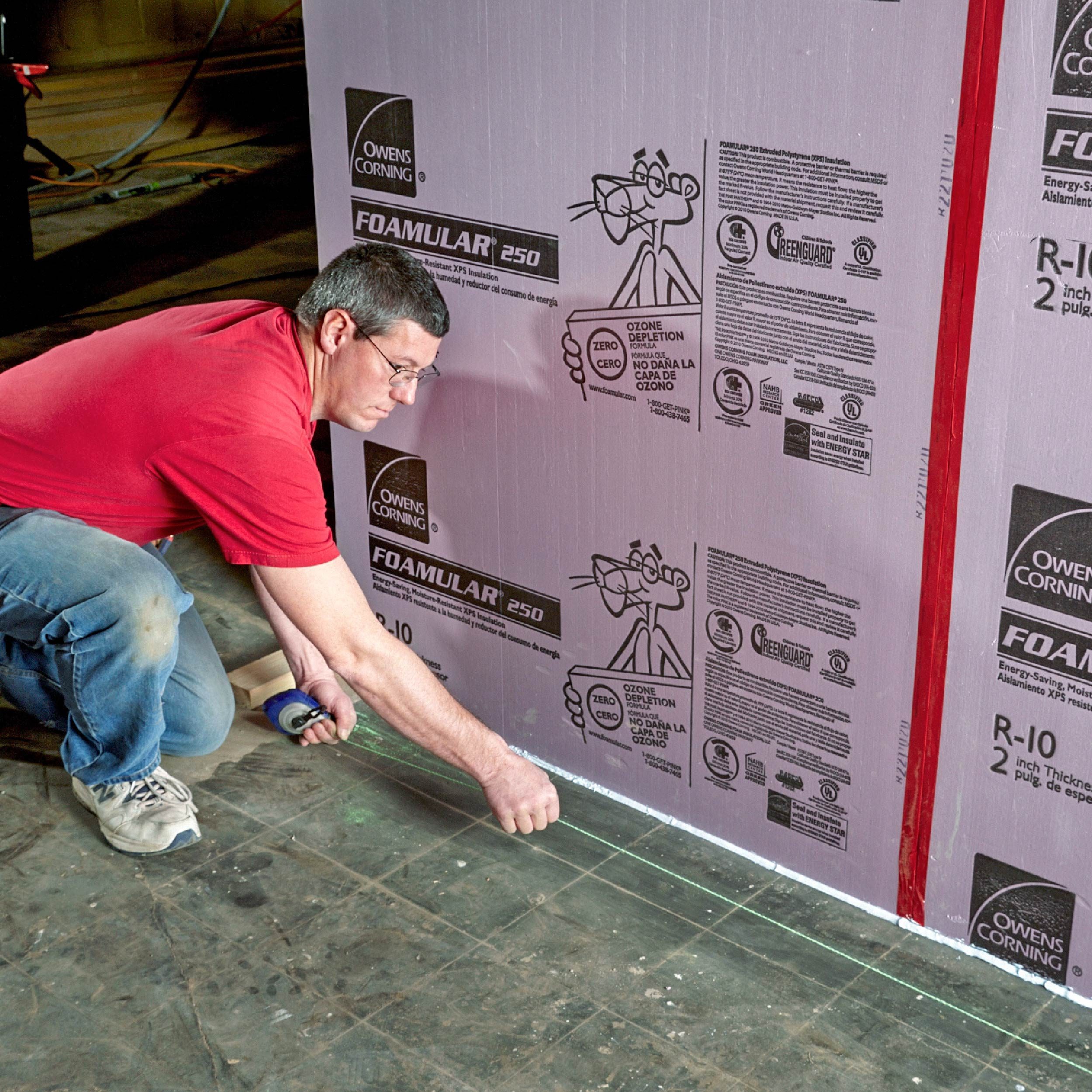 A worker measures and marks a line on the floor next to pink foam insulation boards in a construction environment.
