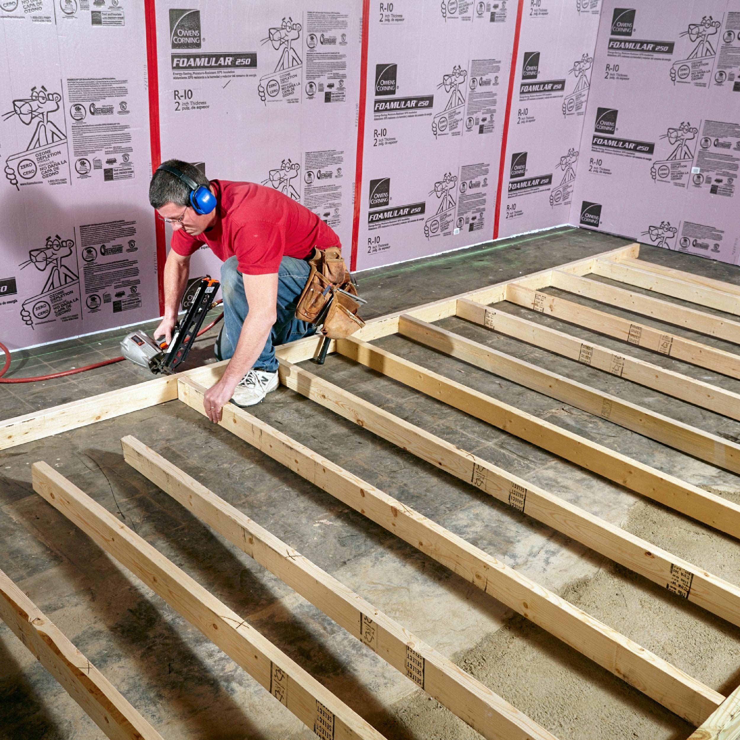 A man in a red shirt secures wooden beams to the floor, surrounded by foam insulation boards in a construction setting.