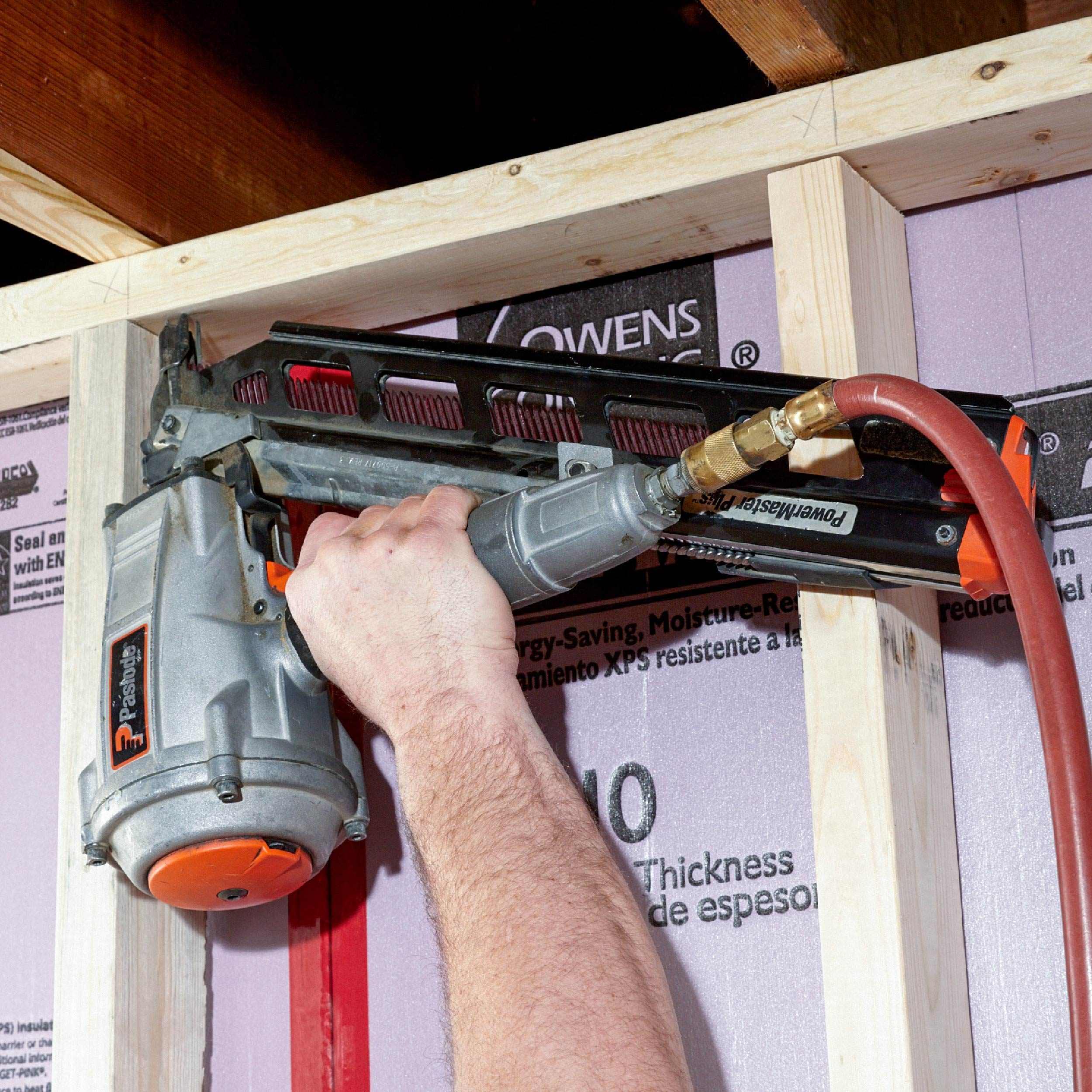 A hand holds a pneumatic nail gun, driving nails into wooden framing in an insulated wall, surrounded by construction materials and framing elements.