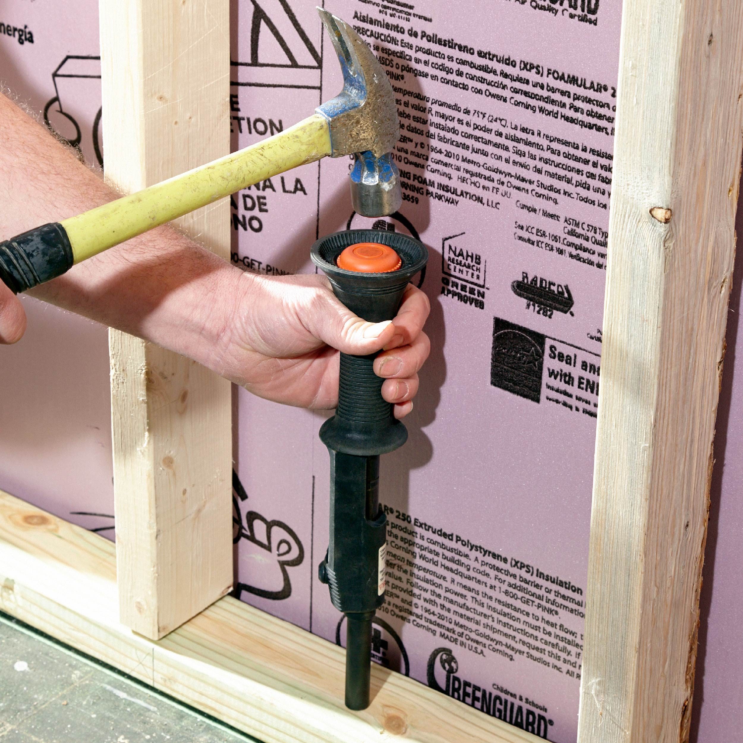 A hand holds a hammer poised to strike a black tool pressing against pink insulation, framed by wooden studs, suggesting a construction or renovation setting.