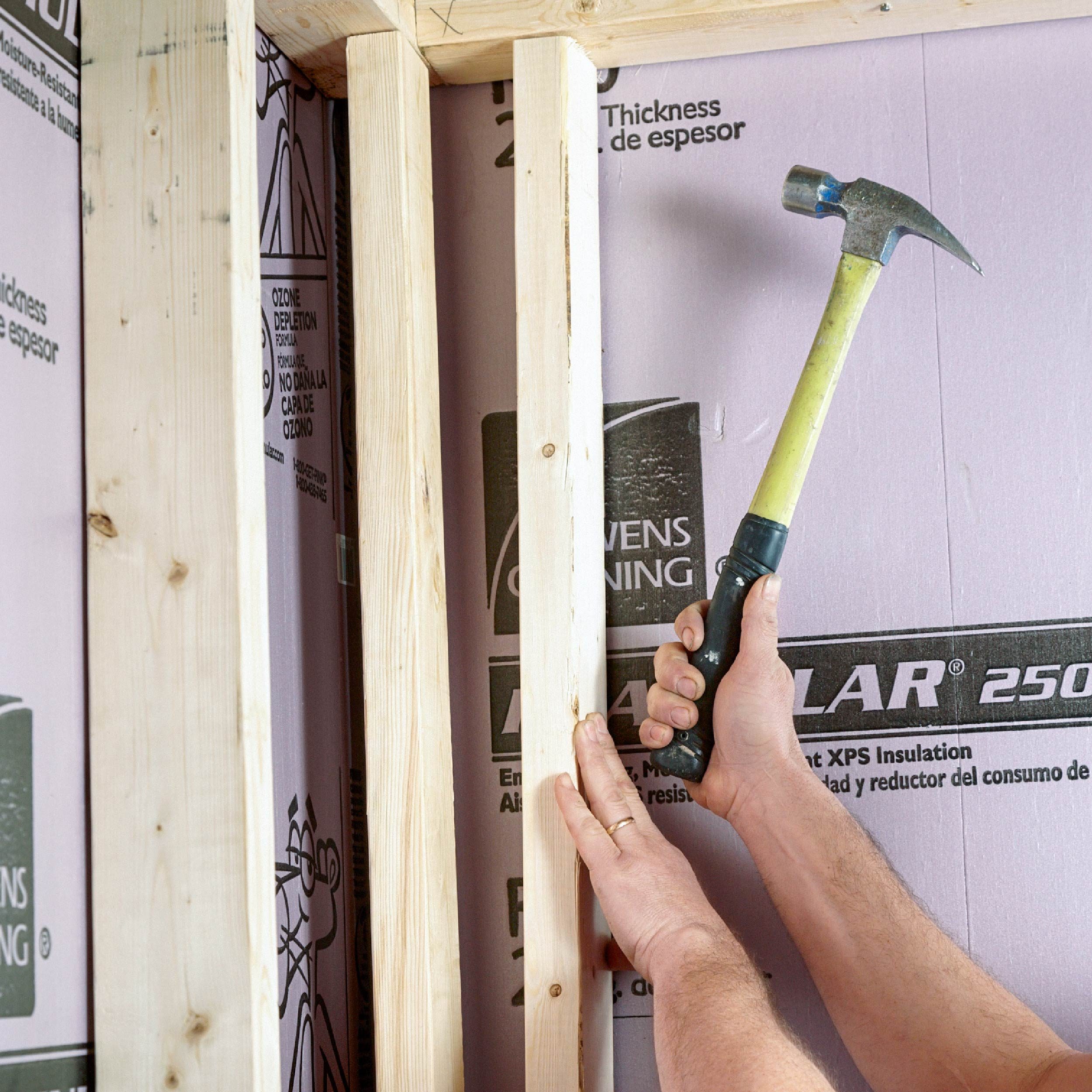 A hand holds a hammer, striking a wooden frame in a construction setting, surrounded by pink insulation panels with various labels.