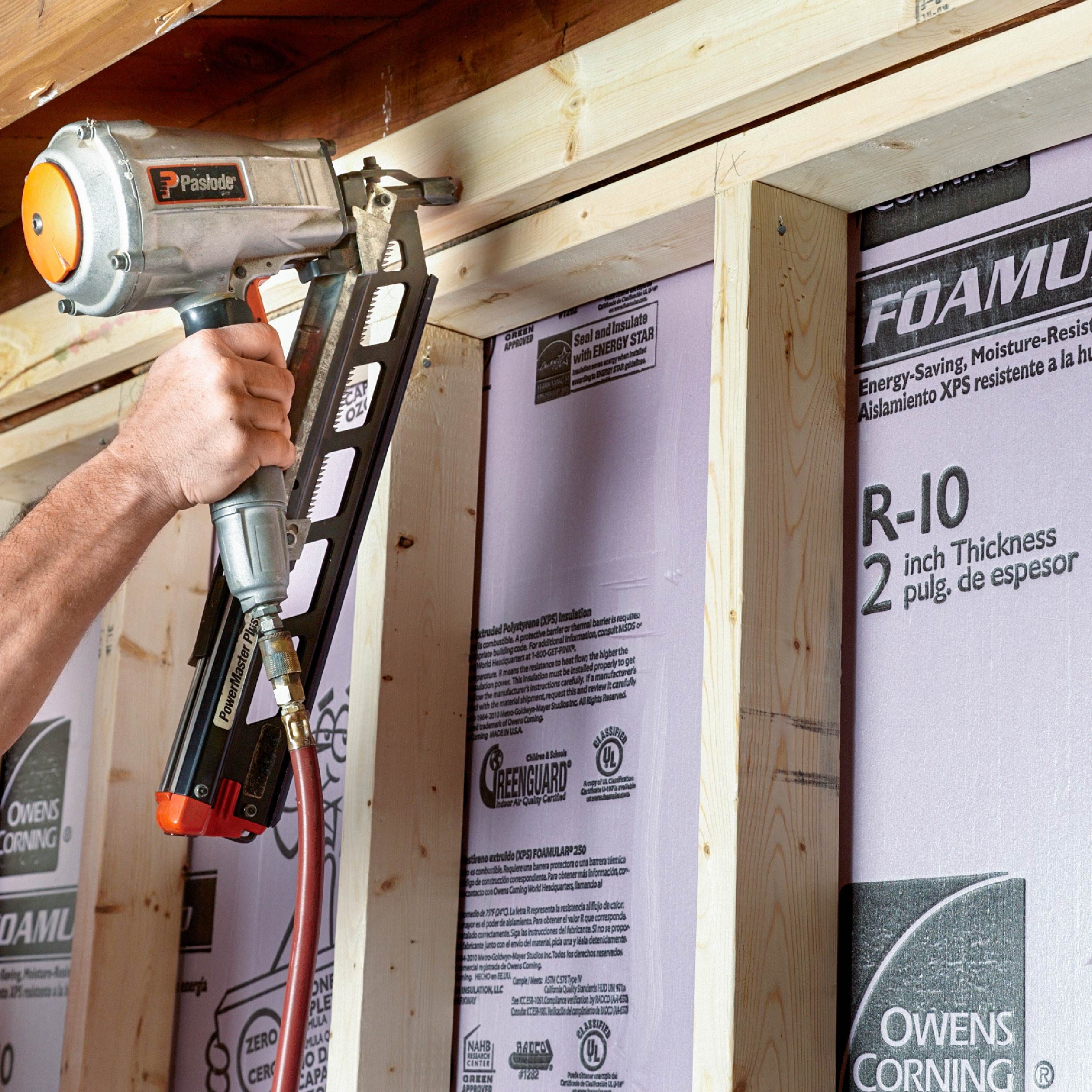 A person uses a nail gun to secure insulation panels between wooden frames in a construction setting, surrounded by wooden structures and insulation materials.