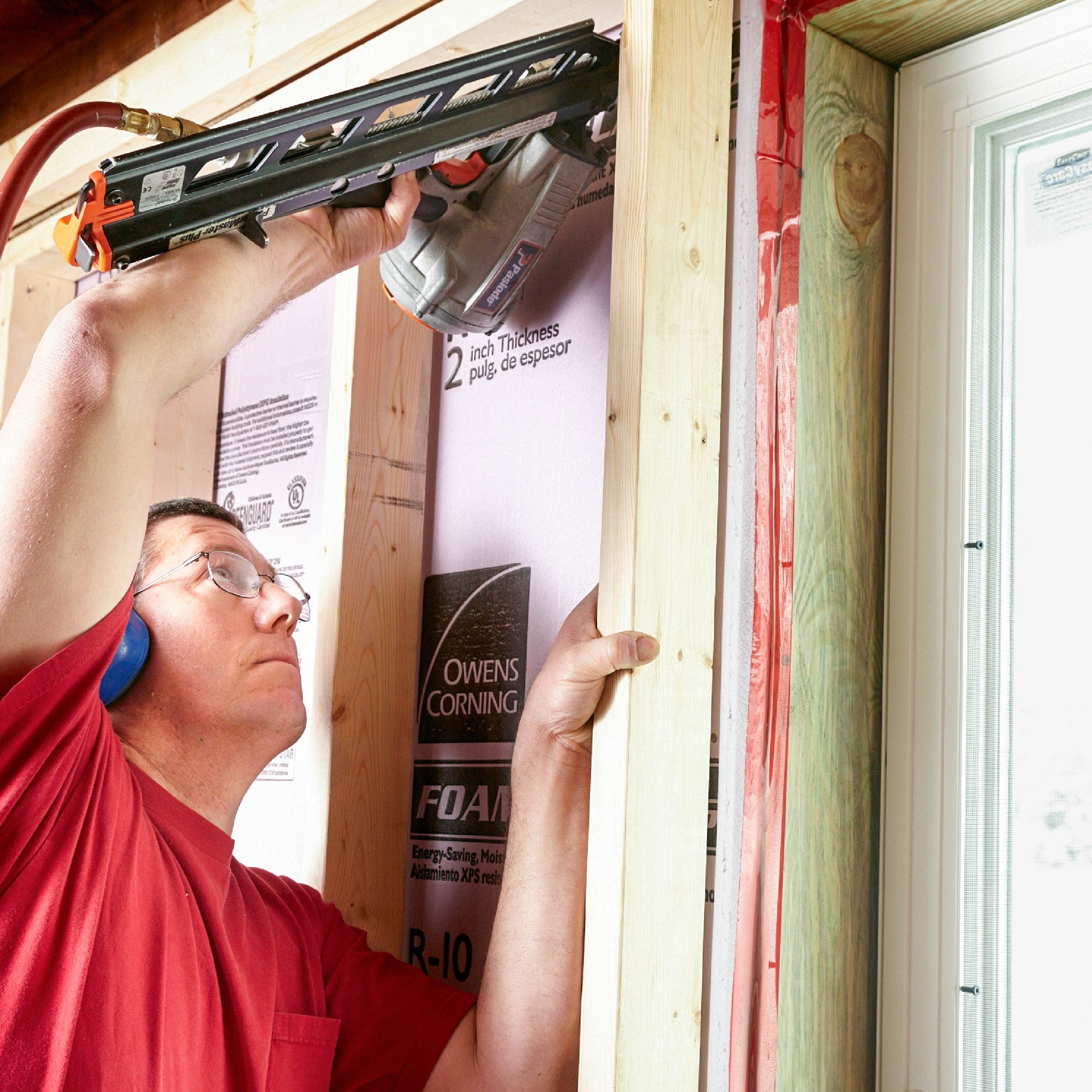 A man uses a power tool to secure insulation between wooden frames in a construction setting, focused on precise placement near a window.
