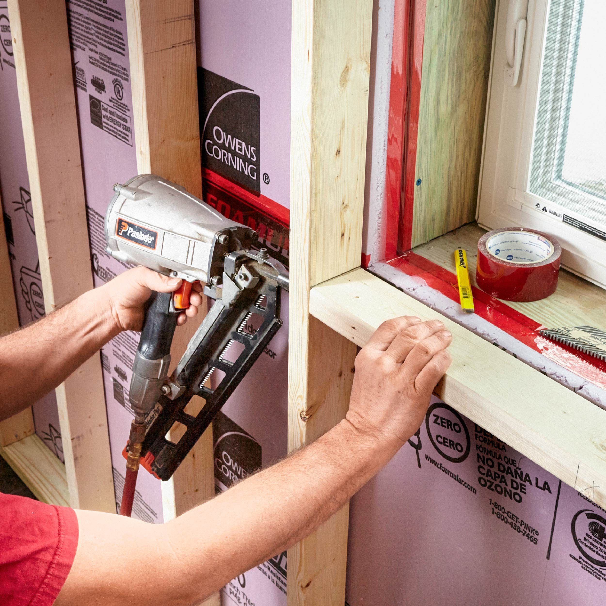 A person uses a nail gun to secure wooden framing around a window in a construction area, with insulation visible on the walls.
