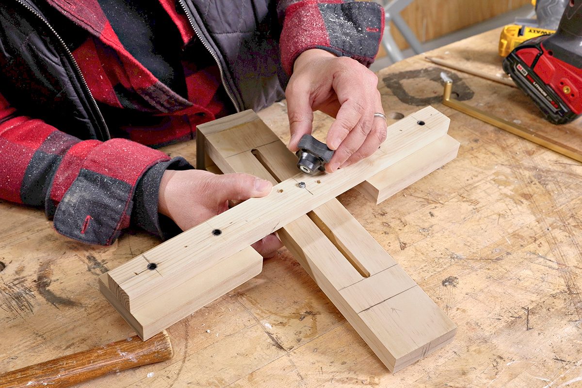 A person in a red plaid shirt uses a small tool to work on a wooden jig clamped together on a workbench, with a drill and other tools nearby.