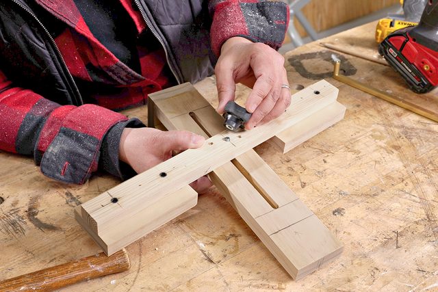 A person in a red plaid shirt uses a small tool to work on a wooden jig clamped together on a workbench, with a drill and other tools nearby.