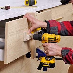 A person uses a yellow power drill to attach a wooden drawer front to a cabinet while holding it steady with a clamp. Other tools are visible on the countertop.