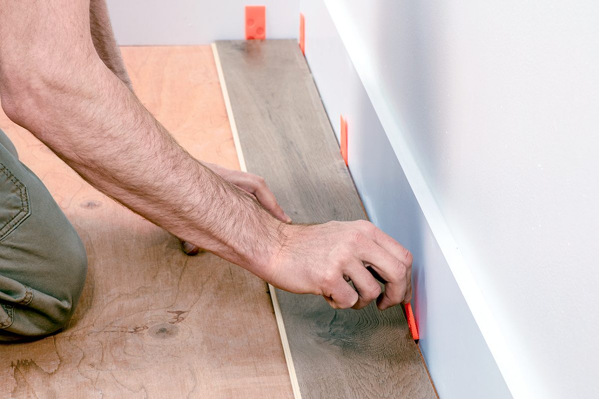 A person installs laminate flooring along a wall, using red plastic spacers to maintain an even gap between the plank and the wall.