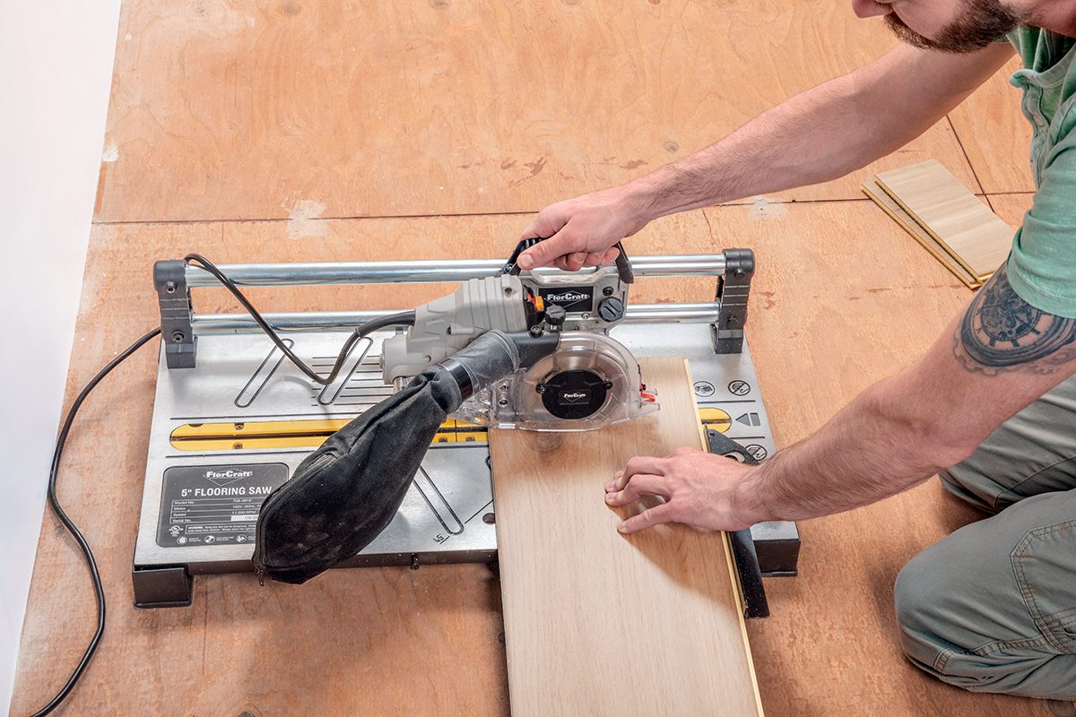 A person using a power saw to cut a piece of wood on a wooden floor; the person’s hands are guiding the wood through the saw