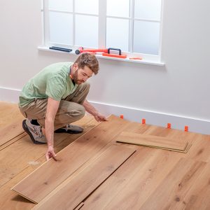 A man in a green shirt and beige pants installs wooden laminate flooring in a bright room with white walls and a window. Tools are placed on the window sill.