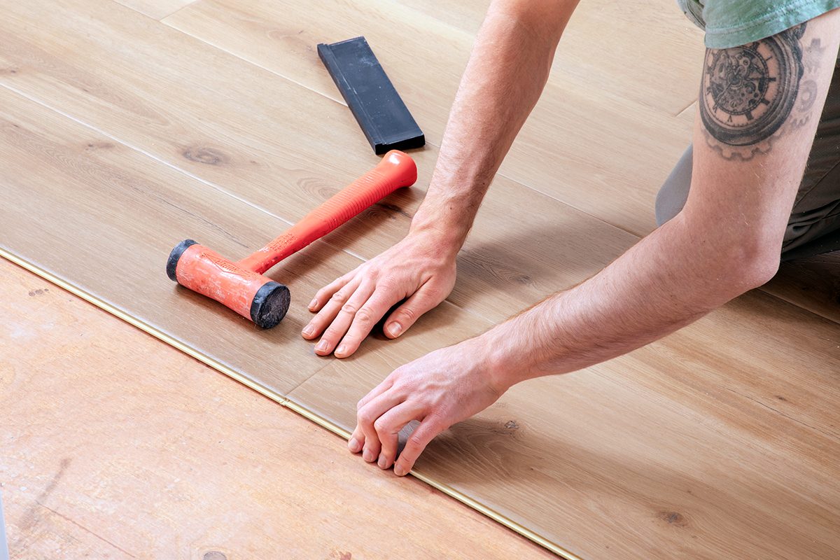 A person installs wooden flooring, using their hands to align the planks. A red rubber mallet and a black tapping block are nearby on the floor. The person’s arm has a visible tattoo.