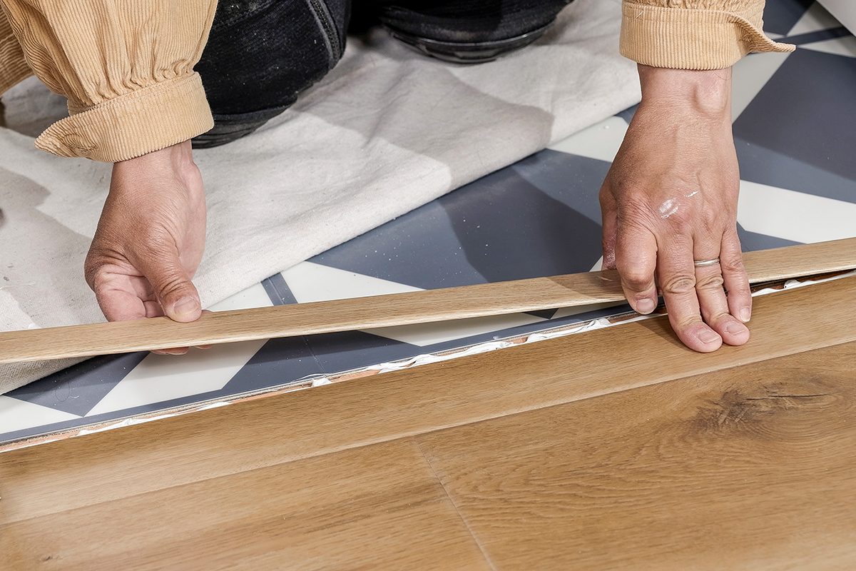 Close-up of a person’s hands installing wood flooring over geometric-patterned underlayment, with a white cloth partially visible in the background.