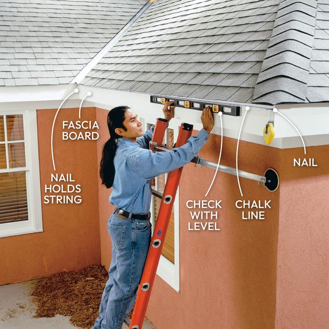 A person uses a level to align a fascia board while standing on a ladder near a sloped roof, with tools and a chalk line visible.