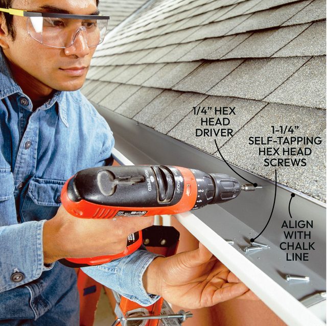 A person is using a power drill to install self-tapping screws into a gutter system on a roof, with shingles in the background.