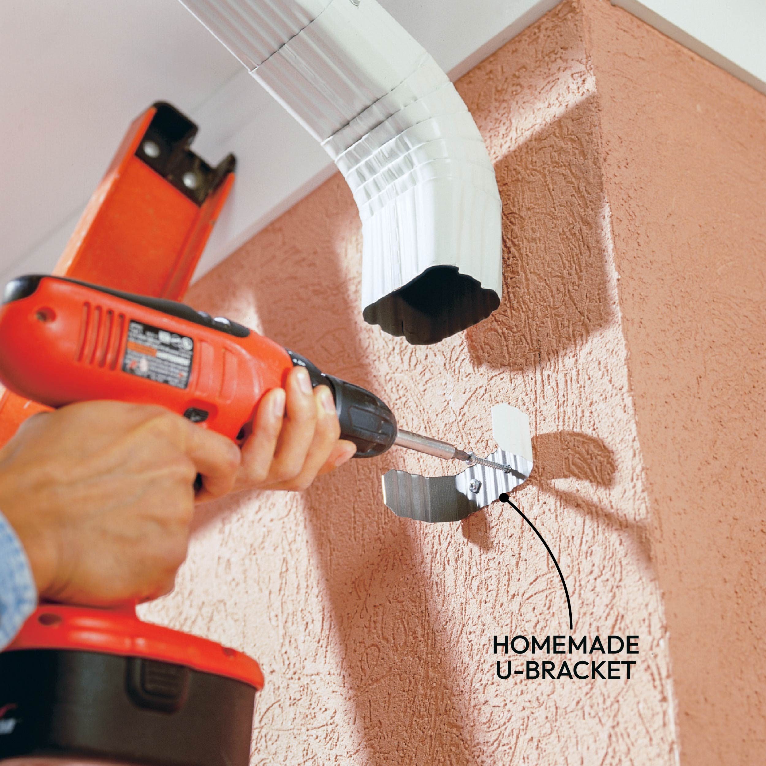A person drills a wall, securing a homemade U-bracket to support a white, curved gutter on a textured pink surface.