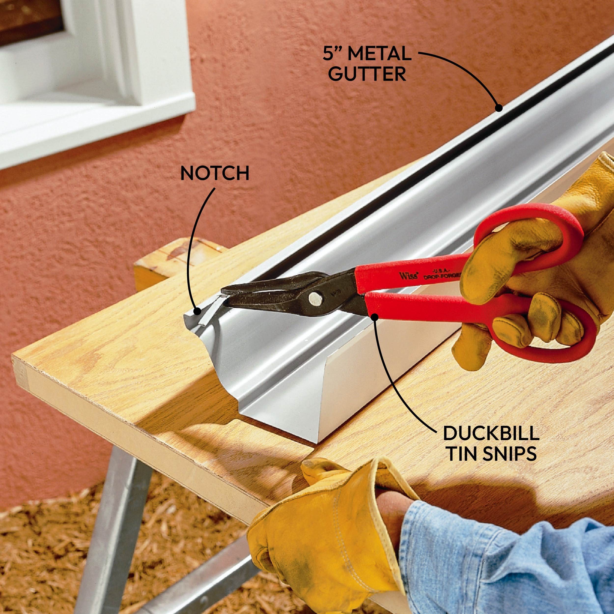 A person uses duckbill tin snips to cut a 5-inch metal gutter with a notch, positioned on a wooden surface, wearing protective gloves.