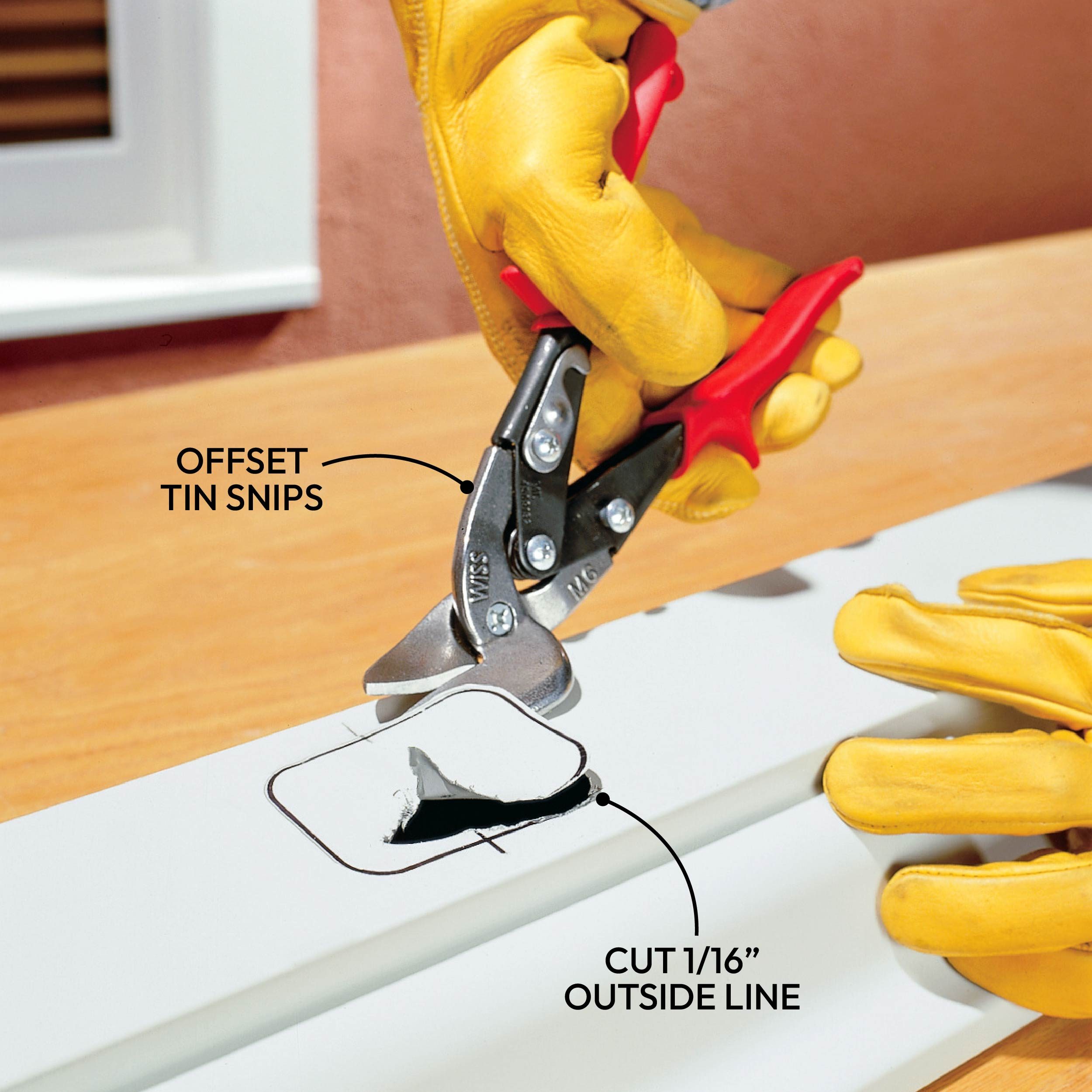 A person uses offset tin snips to cut along a marked line on a white sheet, surrounded by a wooden surface and wearing yellow gloves.