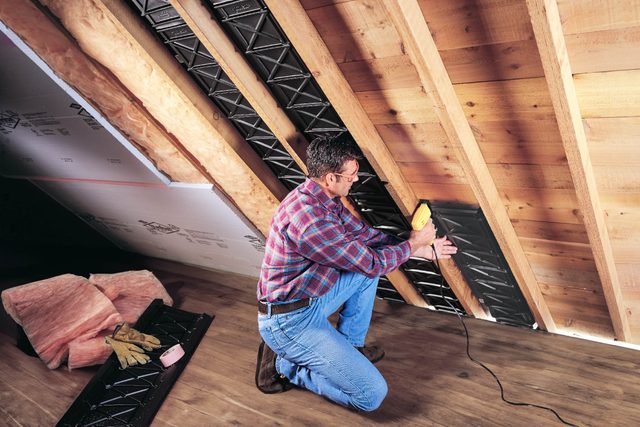 A man kneels while using a power tool to attach materials in an attic with wooden beams and insulation visible in the surroundings.