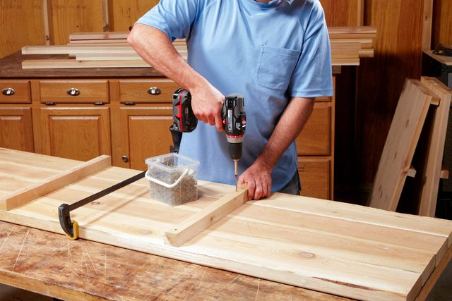 A person drills into a wooden board, securing a piece with a clamp, while a container of screws sits nearby on a workbench.