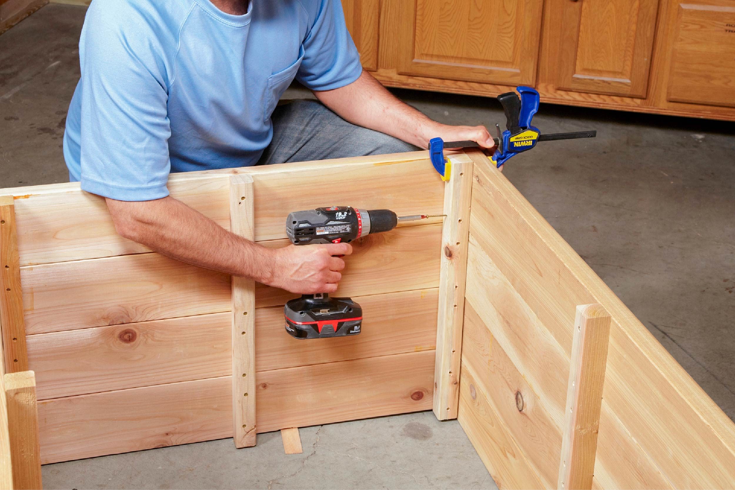 A person uses a power drill to attach wooden panels together, while a clamp holds the pieces in place in a garage setting.