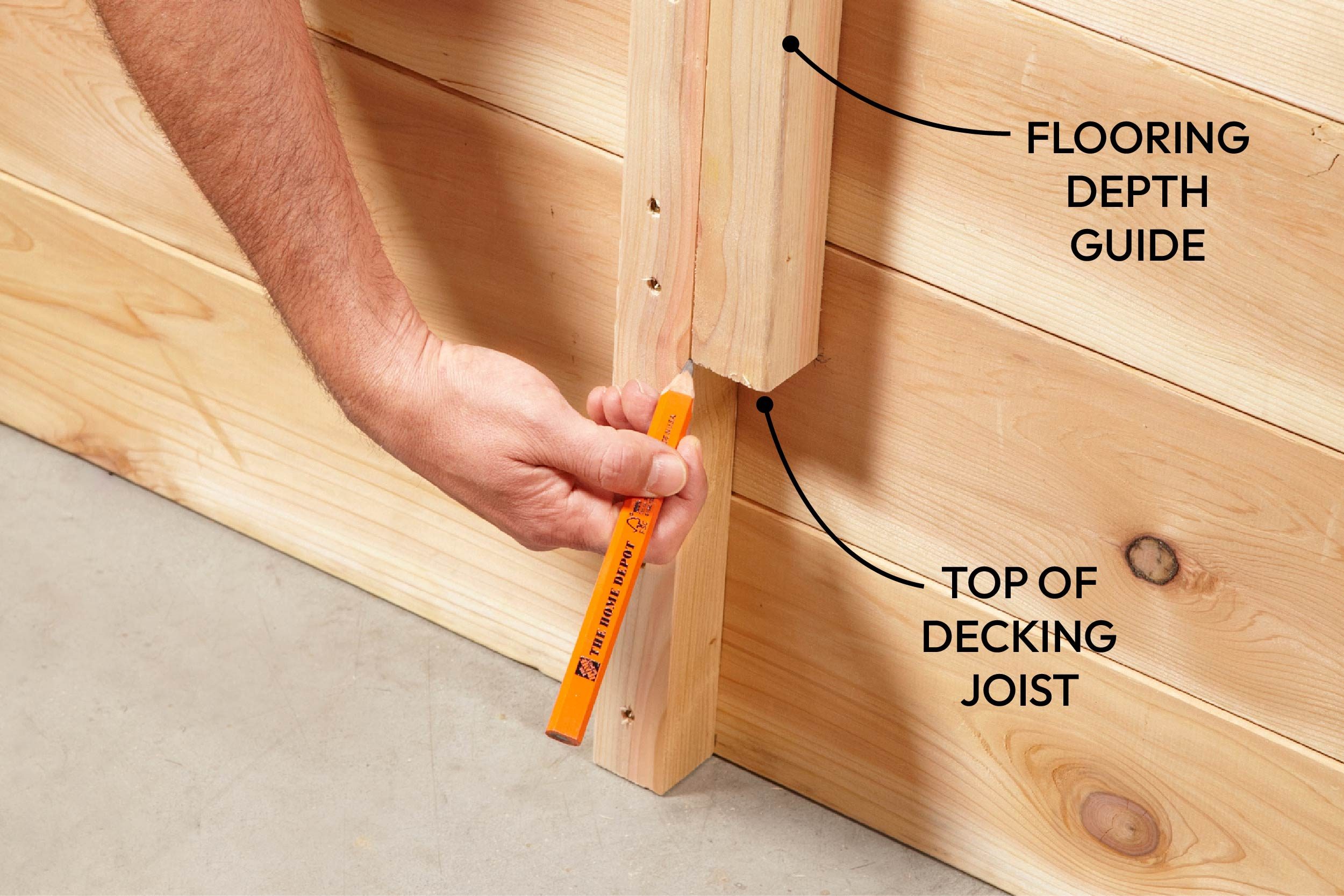 A person holds a pencil against a wooden beam to measure depth, with labeled markings indicating the flooring depth guide and top of the decking joist.