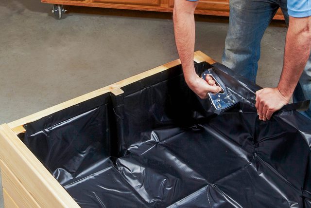 A person attaches a black plastic liner inside a wooden container, working in a garage setting with cabinetry in the background.