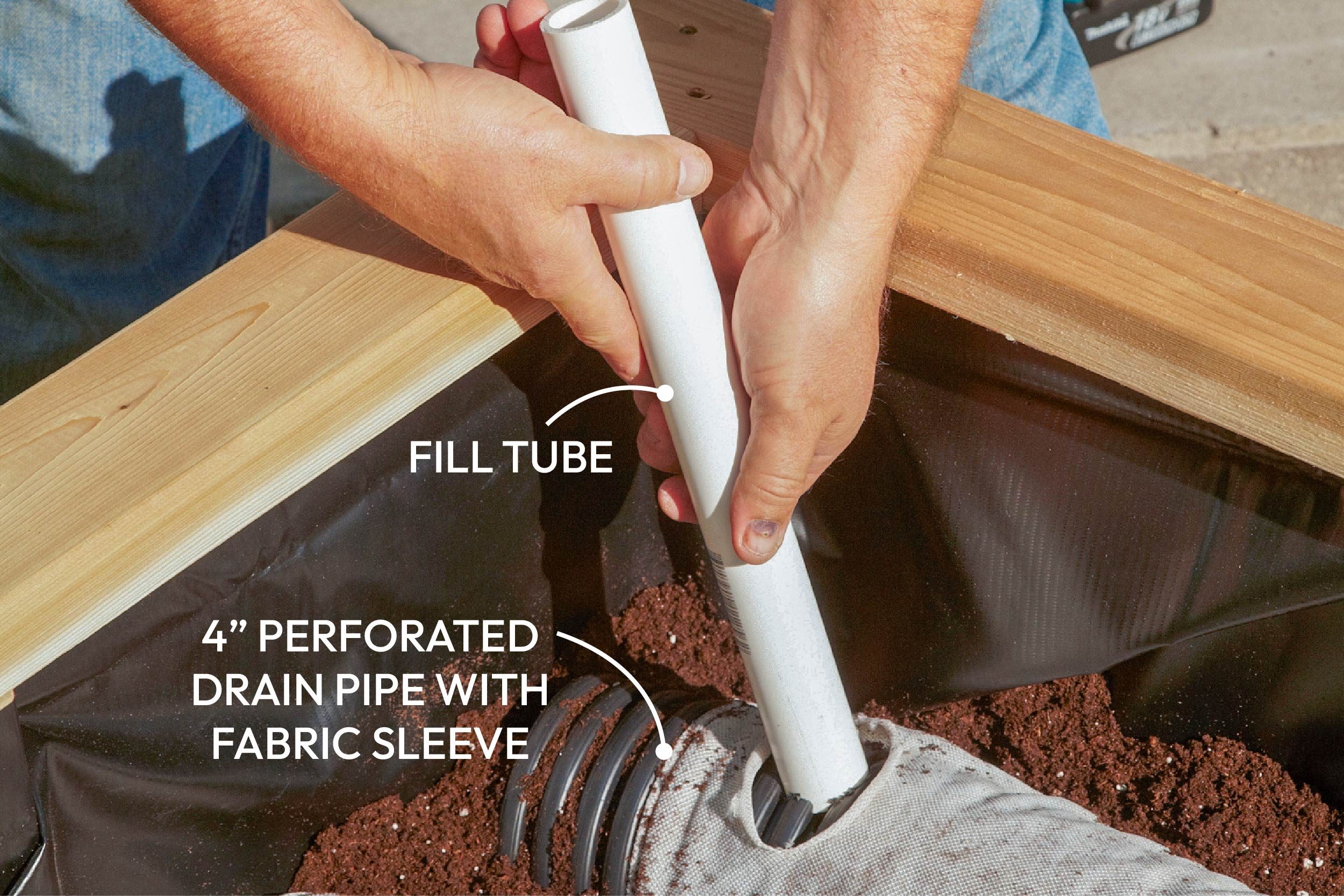A person inserts a white fill tube into a perforated drain pipe placed in soil within a wooden garden bed, under bright sunlight.