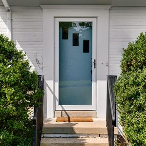 A frosted glass front door with three small clear windows is centered between two green bushes, with black metal railings on each side of the short concrete steps leading up to the entrance.