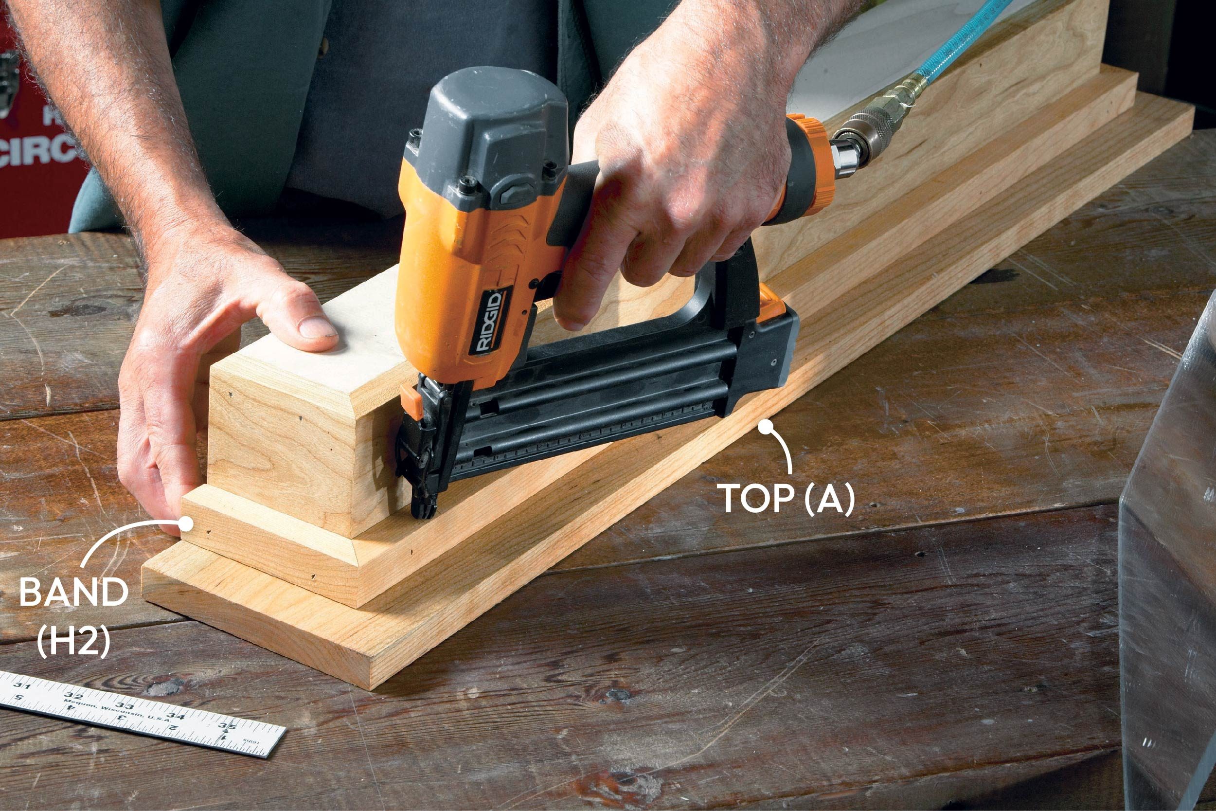 A person uses a nail gun to attach wooden pieces together on a workbench, surrounded by woodworking tools and materials.