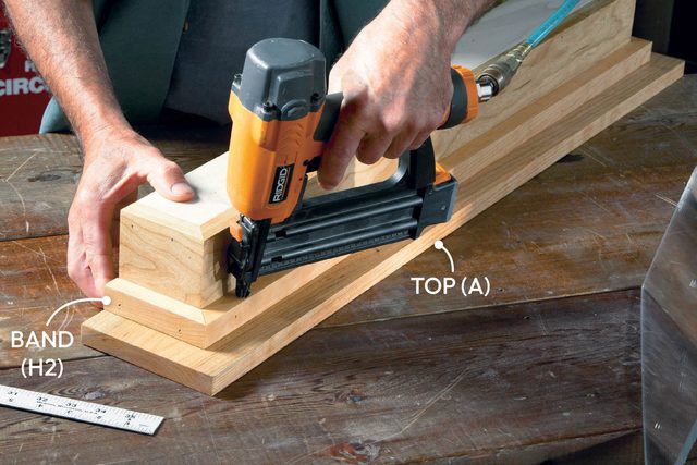 A person uses a nail gun to attach wooden pieces together on a workbench, surrounded by woodworking tools and materials.
