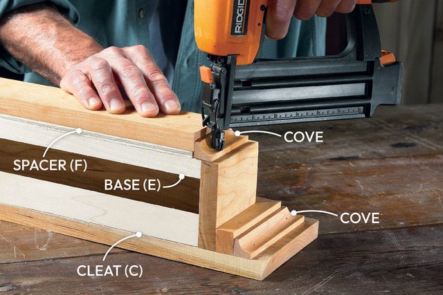 A hand operates a nail gun to attach a wooden base and spacer on a workbench, surrounded by various wooden pieces labeled for assembly.