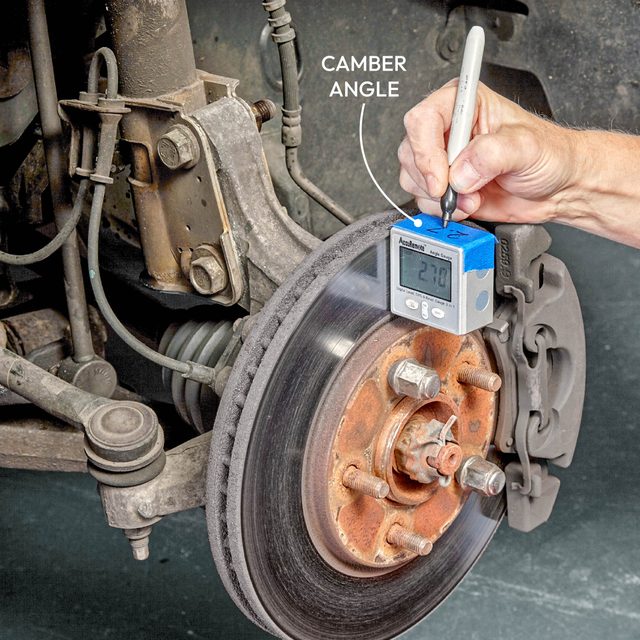 A person measures the camber angle of a vehicle's brake rotor using a digital gauge on a red workshop floor.