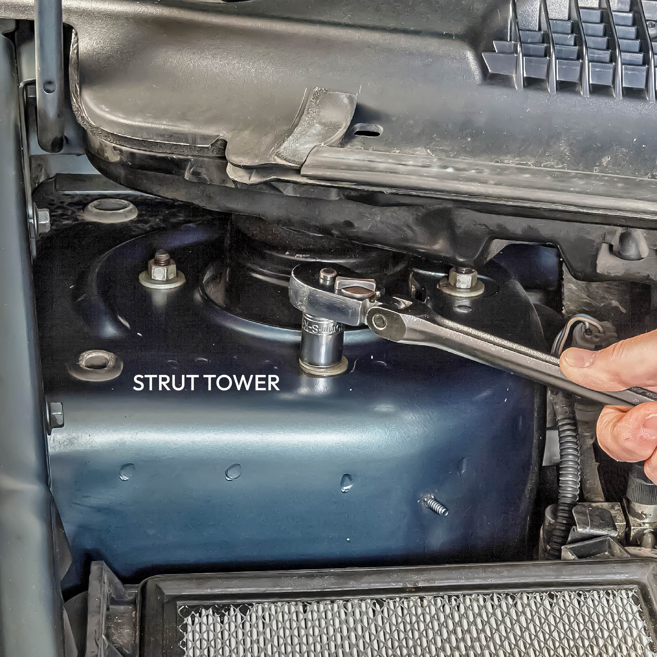 A ratchet wrench fastens a bolt on a strut tower within a vehicle’s engine compartment, surrounded by various mechanical components and fittings.