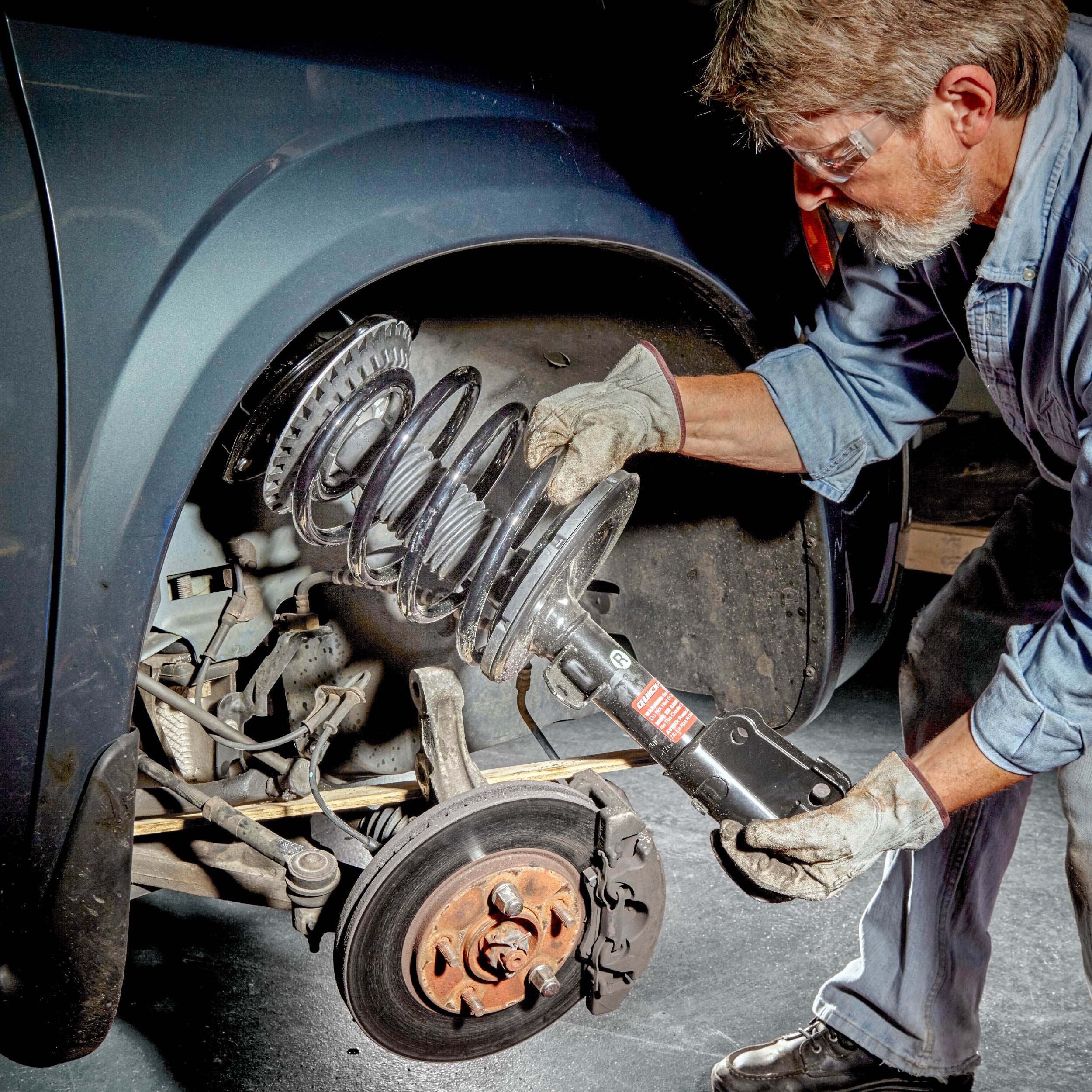 A man is installing a car's suspension component using a tool, surrounded by vehicle parts, in a workshop with a red floor.