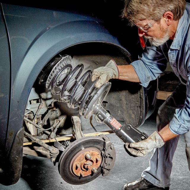 A man is installing a car's suspension component using a tool, surrounded by vehicle parts, in a workshop with a red floor.