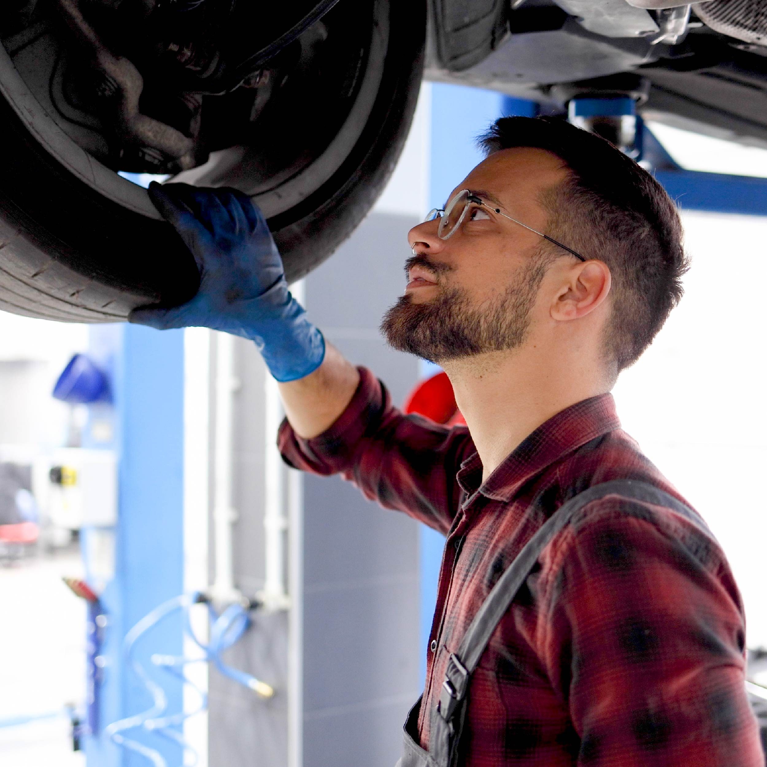 A mechanic inspects a vehicle's undercarriage, in a well-lit garage with tools and equipment visible in the background.
