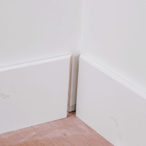 Close shot of two white baseboards meet at an inside corner against a white wall; with a small gap visible between them; The floor is light-colored wood;