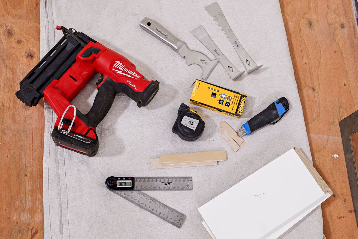 An overhead shot displays essential carpentry tools arranged on a clean white mat placed over a wooden floor; The collection includes a vibrant red nail gun for fast fastening tasks; a precise tape measure and ruler for accurate measurements; handy scraper tools and a utility knife for cutting and shaping materials; a digital angle finder for achieving perfect angles; sturdy wood strips prepared for assembly; and an open box filled with nails;